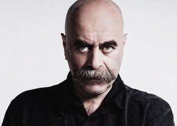 siyah giyimli, kel ve bıyıklı bir adam objektife bakıyor