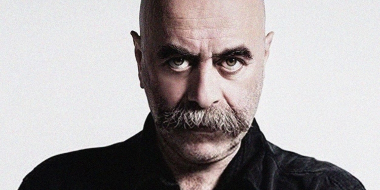 siyah giyimli, kel ve bıyıklı bir adam objektife bakıyor