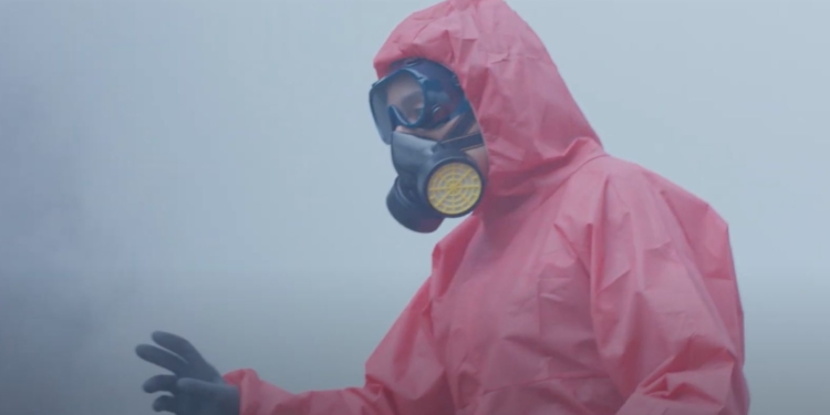 Tam korumalı giyinmiş adam bir sisin içinde elini yana doğru kaldırıyor. Üstünde koruma giysisi ve yüzünde de maske var.