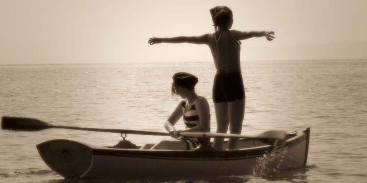 İki çocuk bir sandalda. Biri kollarını açmış ayakta, diğeri de sandalda oturuuyor.