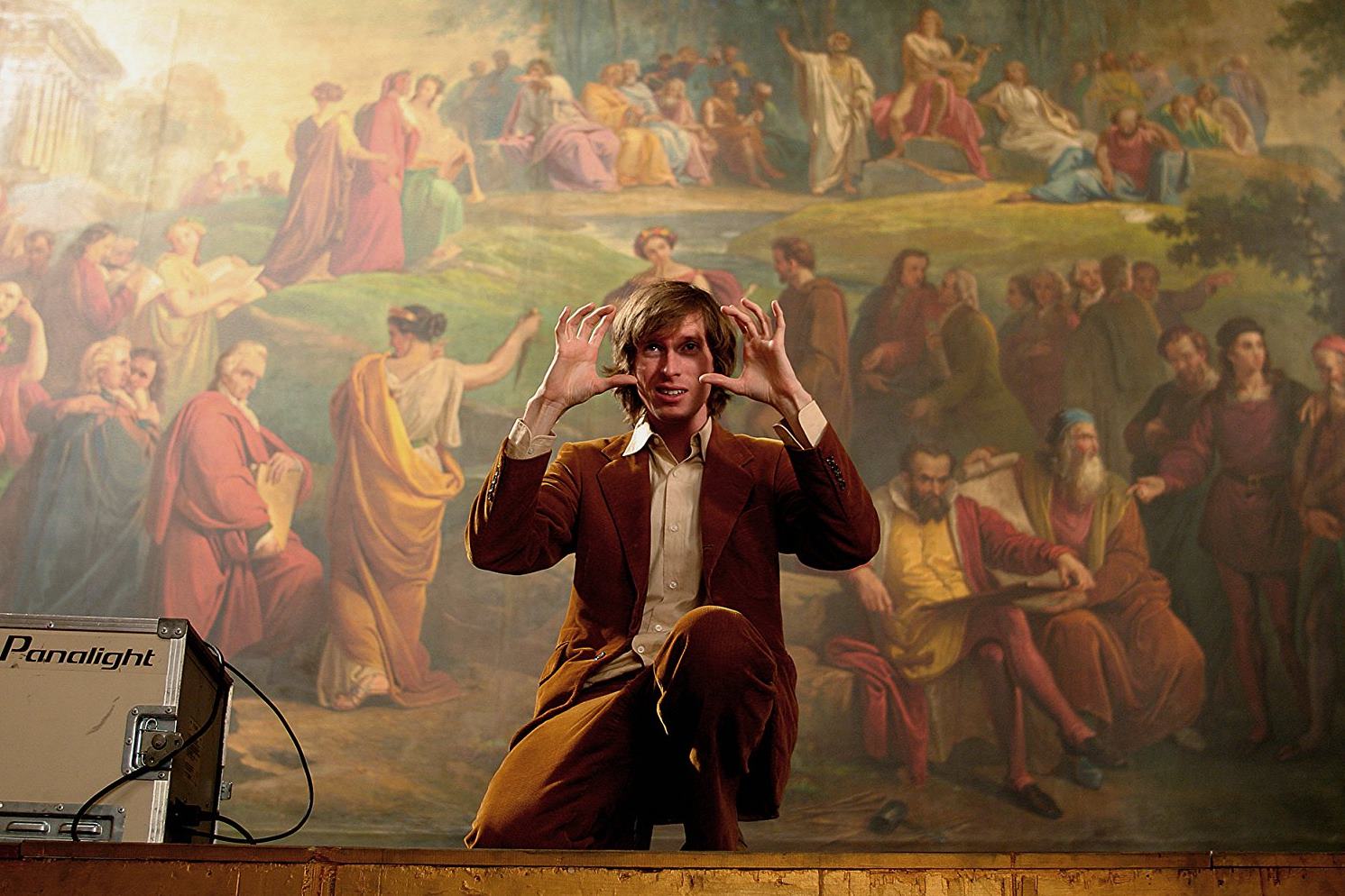 Bir adam, bir tablonun önünde durup elleriyle kamera işareti yapıyor.
