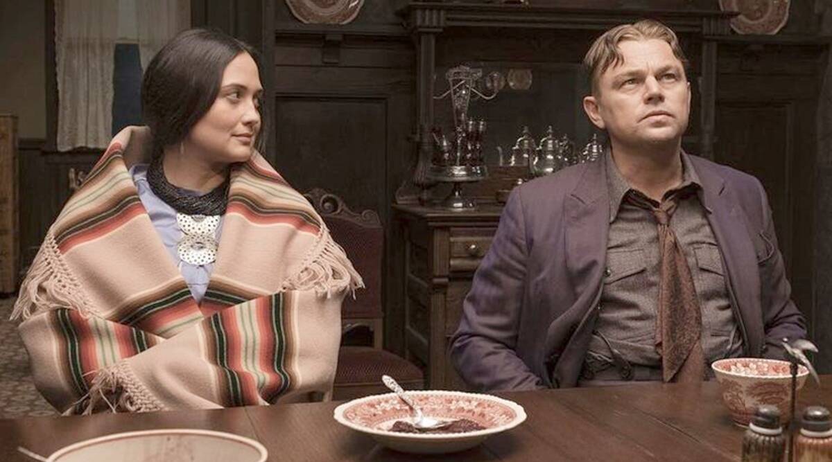 Bir kadın ve adam yemek masasının etrafında oturuyor. Kadının üstünde panço var, adam takım elbiseli.